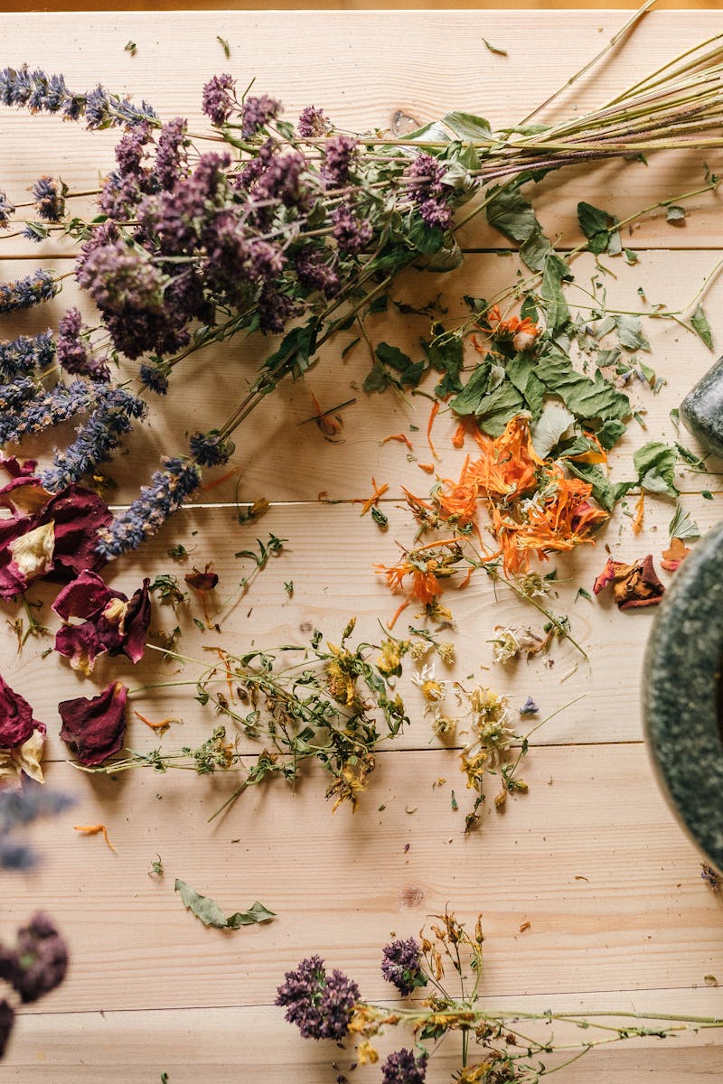 Flat lay of dried herbs and flowers with a stone mortar and pestle on a rustic wooden table.